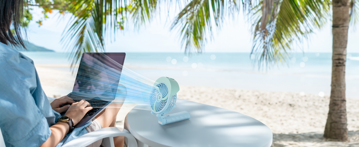Person using laptop on beach chair with small portable fan nearby. Palm trees and ocean visible in background.
