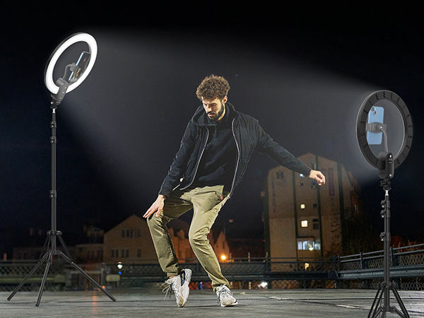Two ring lights on stands illuminate a person dancing outdoors at night. The lights feature adjustable mounts for phones or cameras.