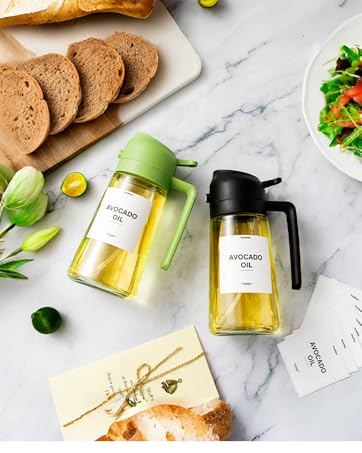 Two glass oil dispensers with pump tops, one green and one black, next to bread slices and a salad on a marble surface.