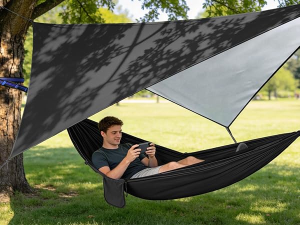 Outdoor hammock setup with black fabric hammock suspended between trees, with gray tarp canopy providing shade overhead.
