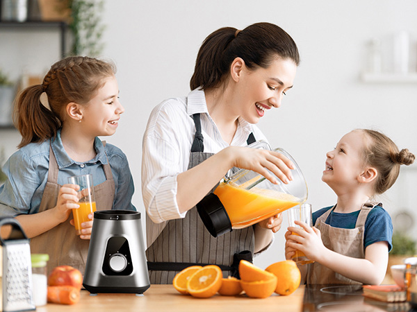 Stainless steel blender with pitcher on kitchen counter. Woman pours orange juice from pitcher into child's glass. Fresh oranges and other fruits nearby.