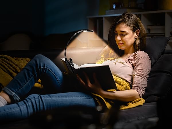 Person sitting on a couch in dimly lit room, reading a book with a small lamp providing focused light.