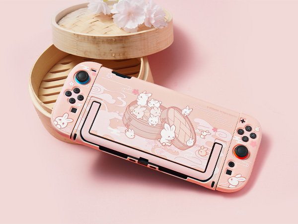 Pink Nintendo Switch gaming console with decorative cat-themed protective case, displayed on pink background with circular bamboo container.