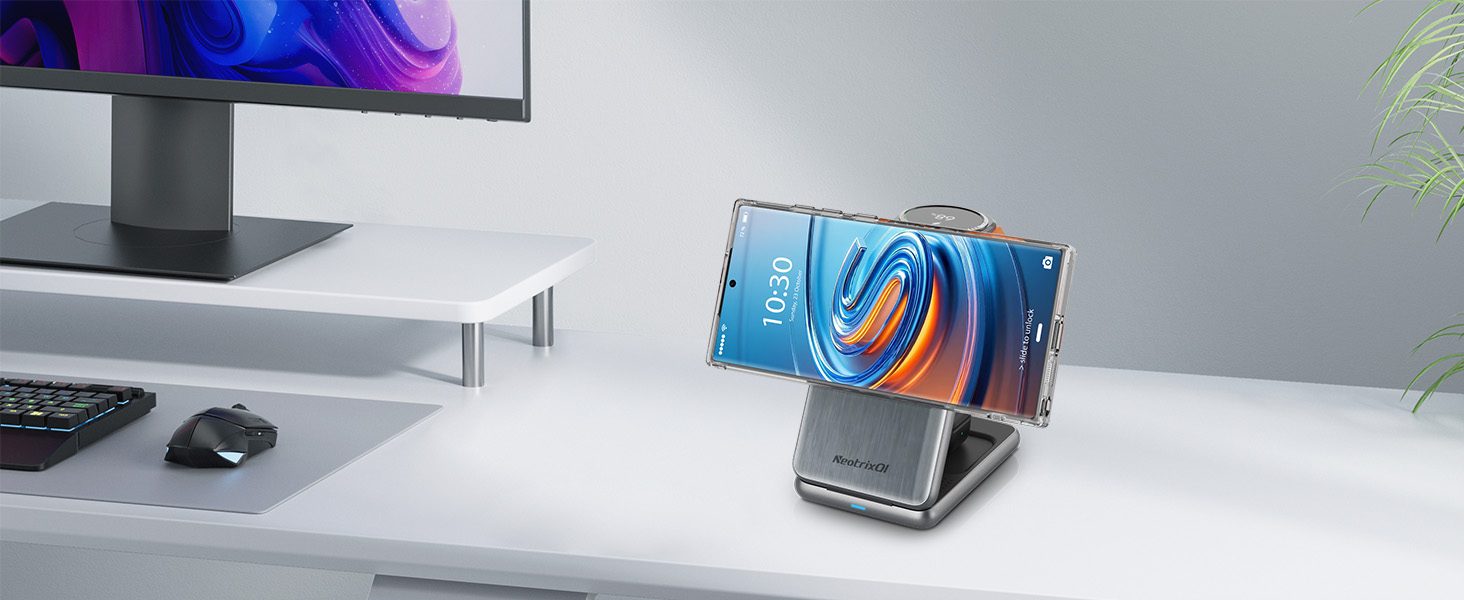 Smartphone displayed on a metal stand, showing colorful swirling wallpaper. Positioned on white desk with computer monitor, keyboard, and mouse visible in background.