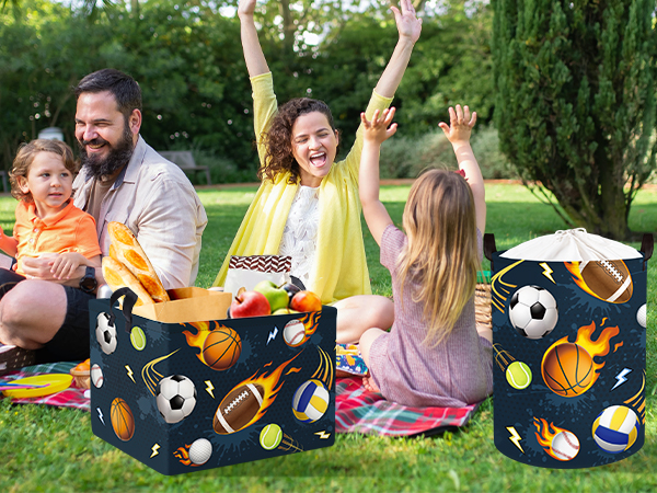 Two navy blue storage bins with sports-themed pattern featuring footballs, basketballs, and baseballs. People enjoying outdoor picnic on grass with storage containers visible.