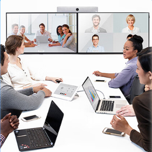 Business meeting scene with people in an office and on video screens. Participants are using laptops and engaging in discussion across physical and virtual spaces.