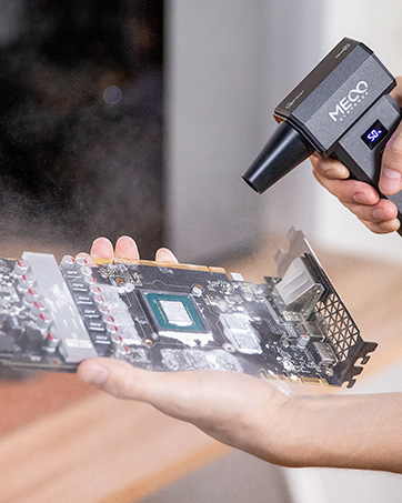 Handheld air blower cleaning a computer graphics card. Device labeled 'XPRO' sprays compressed air onto circuit board components, removing dust particles visible in the air.