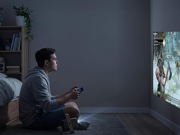 Compact projector displaying a vibrant forest scene on a wall. Person sits on floor holding game controller, suggesting interactive gaming or entertainment use.