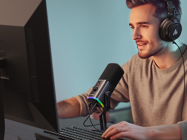Black USB condenser microphone with RGB lighting, positioned in front of a computer user wearing headphones, suggesting use for podcasting or streaming.