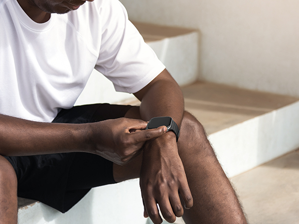 Person seated on white couch looking down at digital device or smartwatch on wrist, wearing casual white t-shirt and dark shorts.