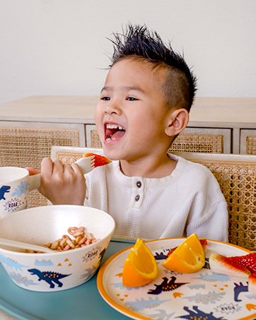 Boy smiling while eating fruit from zak! designs Dino 3-piece dinnerware set