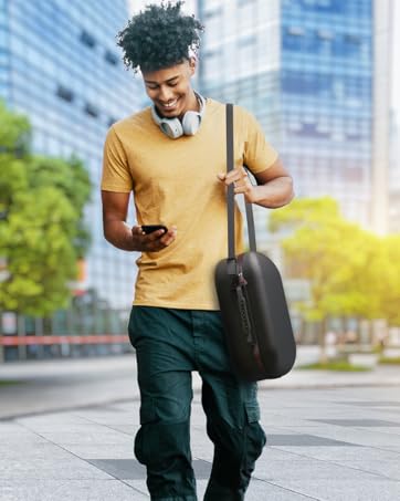 Man in yellow shirt walking on city street, carrying shoulder bag and using smartphone. Headphones around neck, buildings and trees in background.