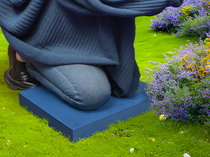 Person kneeling on a blue foam pad while gardening. Surrounded by colorful flowers and green lawn, demonstrating comfortable outdoor work posture.