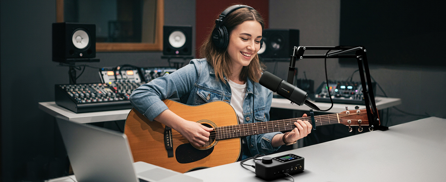 Professional recording studio setup with acoustic guitar, microphone on boom arm, audio interface, and studio monitors on desk against wood-paneled wall.