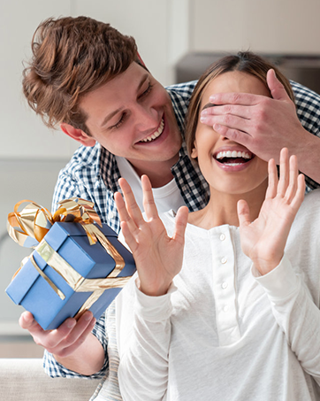 Blue gift box with gold ribbon and bow. Person holding the box while surprising another person, covering their eyes in a playful gesture.