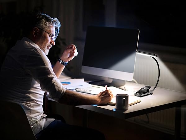 Person working at a desk in a dark room, illuminated by a computer screen and desk lamp. They appear to be writing or drawing.