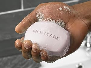 Close up of hand holding lathered Dove Men+Care cleansing bar with faucet and tiles in background.