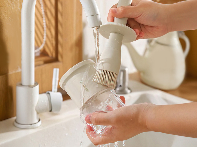 A hands-free soap dispenser brush attachment for faucets. Water flows through the brush head, mixing with soap to clean dishes. Demonstrated in use over a sink.