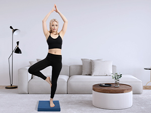 Woman in black athletic wear performing yoga tree pose on blue mat in living room with couch, coffee table, and floor lamp.