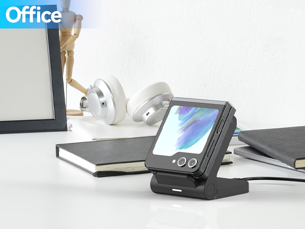 Portable electronic device with folding stand on office desk, displaying colorful screen. White headphones and books visible nearby.