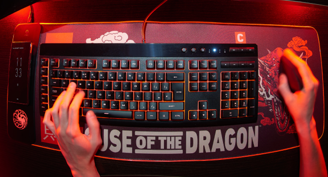 Gaming keyboard with red backlighting and hands positioned for typing. Text 'ISE OF THE DRAGON' visible on the keyboard or surrounding area.