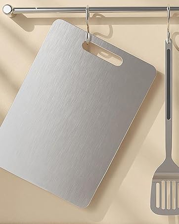 Stainless steel kitchen utensils hanging on a rod. Large rectangular cutting board with handle and a slotted spatula visible. Brushed metal finish on both items.
