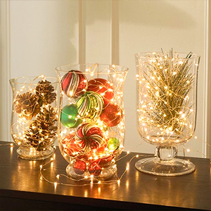 Three glass containers filled with decorative fairy lights. One contains pinecones, another colorful ornaments, and the third has a miniature tree, creating a warm ambiance.