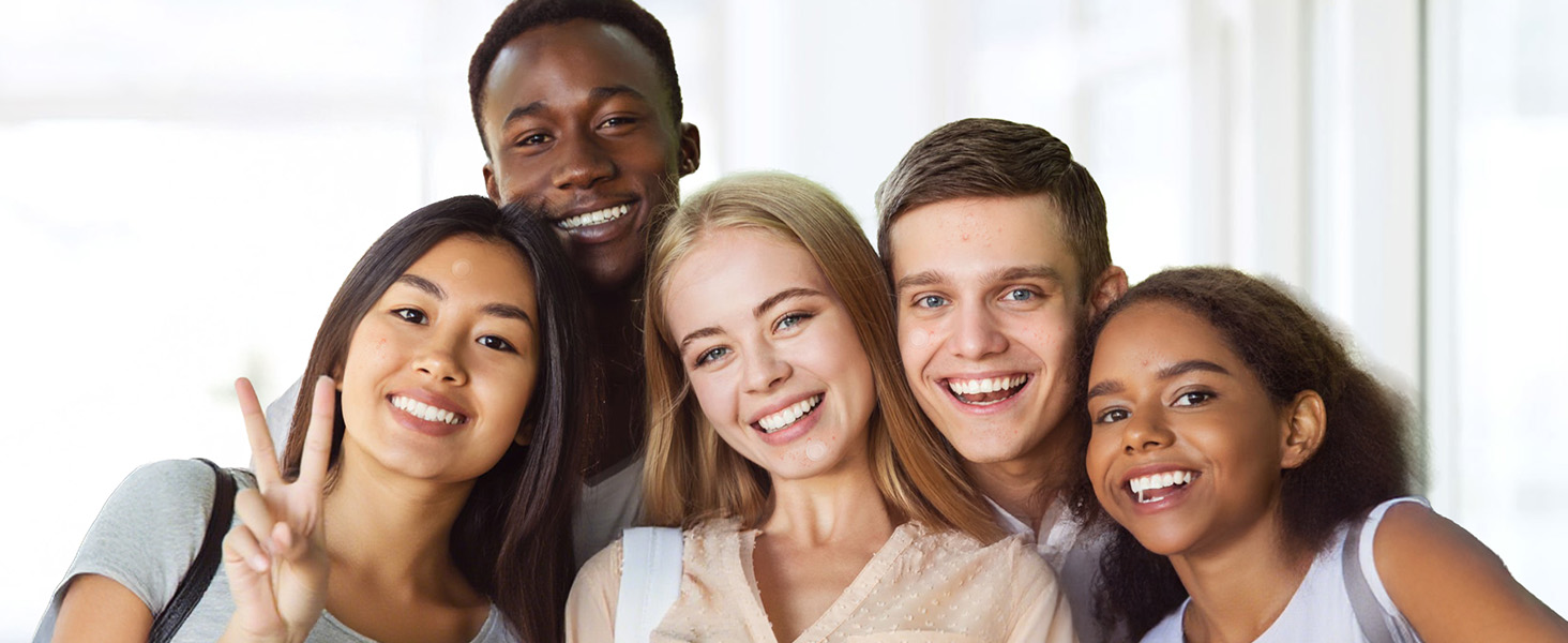 Five diverse young adults smiling and posing together indoors against a bright white background, appearing happy and relaxed in casual clothing.