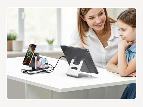 Wireless charging stand with multiple device support on a desk. Adjacent tablet stand holds device upright. Two people observe the setup in a bright room.