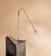 Copper-colored bent wire with loop at top and hook at bottom, balancing on edge of rough concrete or stone block.