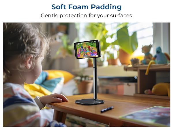 Tablet stand with soft foam padding protection, shown in use on wooden desk with indoor plants in background.