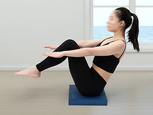 Woman performing a V-sit exercise on a blue yoga mat, demonstrating core strength and flexibility in a bright indoor setting.