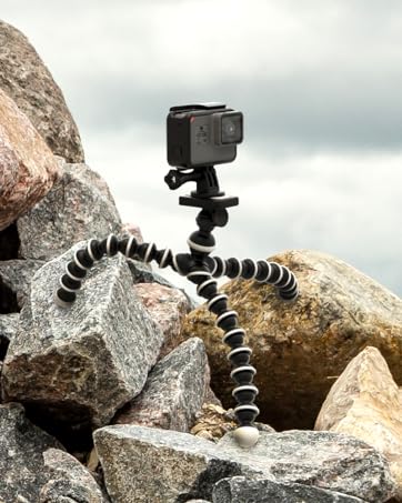 Black action camera mounted on flexible tripod with segmented legs. Set up on rocky terrain, likely for outdoor filming or photography.