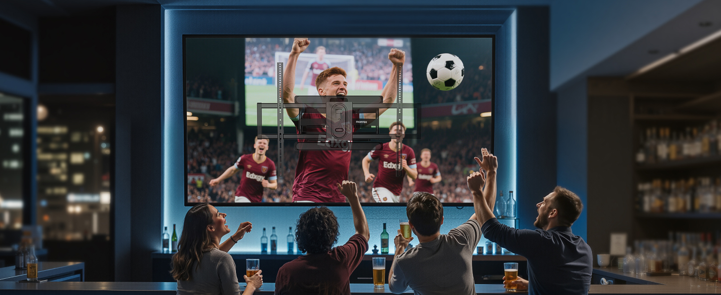 Large screen displaying a soccer match with players visible. Several people in foreground watching the screen in a dimly lit room.