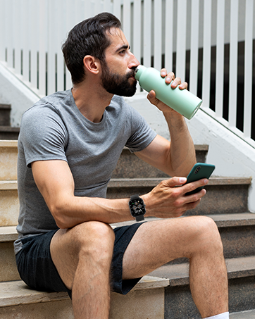 Man sitting on stairs drinking from a light green insulated water bottle. He's wearing gray t-shirt, shorts, and holding a smartphone.