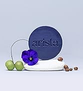 Circular navy blue object with 'arista' embossed, displayed with a purple flower, green apples, and coffee beans on a white surface.
