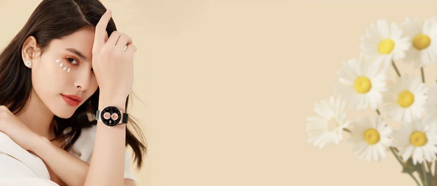 Circular smartwatch with black strap and display showing a cartoon skull face. Woman's wrist modeling the watch against a beige background with white daisies.