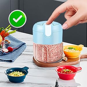 Light blue electric food chopper processing pink contents, with small colorful bowls and citrus fruit visible on wooden surface.