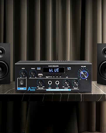 Audio amplifier with digital display, multiple inputs, and control knobs, flanked by two black bookshelf speakers. System sits on marble surface against striped curtain background.