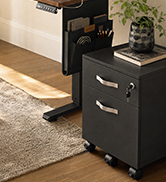 Black bedside nightstand with two front drawers and metal handles, positioned on wooden flooring next to bed with green plant visible in background.