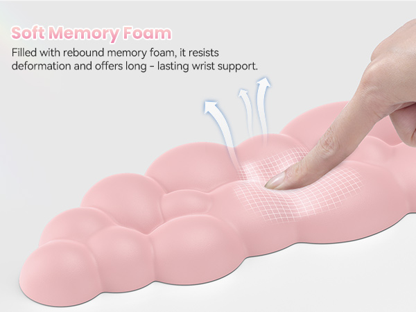 Pink ergonomic wrist rest made of soft memory foam. Finger presses into cushioned surface, demonstrating rebound support. Textured, cloud-shaped design for long-lasting comfort.