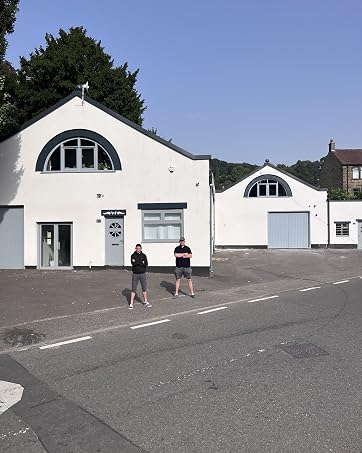 Two white buildings with arched windows, possibly warehouses or workshops. The buildings have large doors and are situated on a paved area with trees visible in the background.