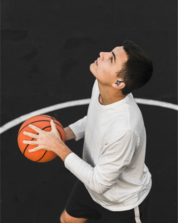 Person in white long-sleeved shirt holding an orange basketball, looking upwards against a black background. Partial view of a white circular line visible.