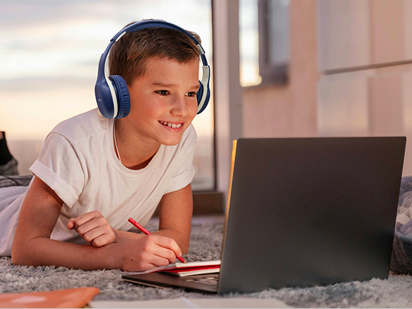 Blue over-ear headphones worn by a child using a laptop on a carpeted floor. The headphones have a padded headband and ear cups for comfort during extended use.