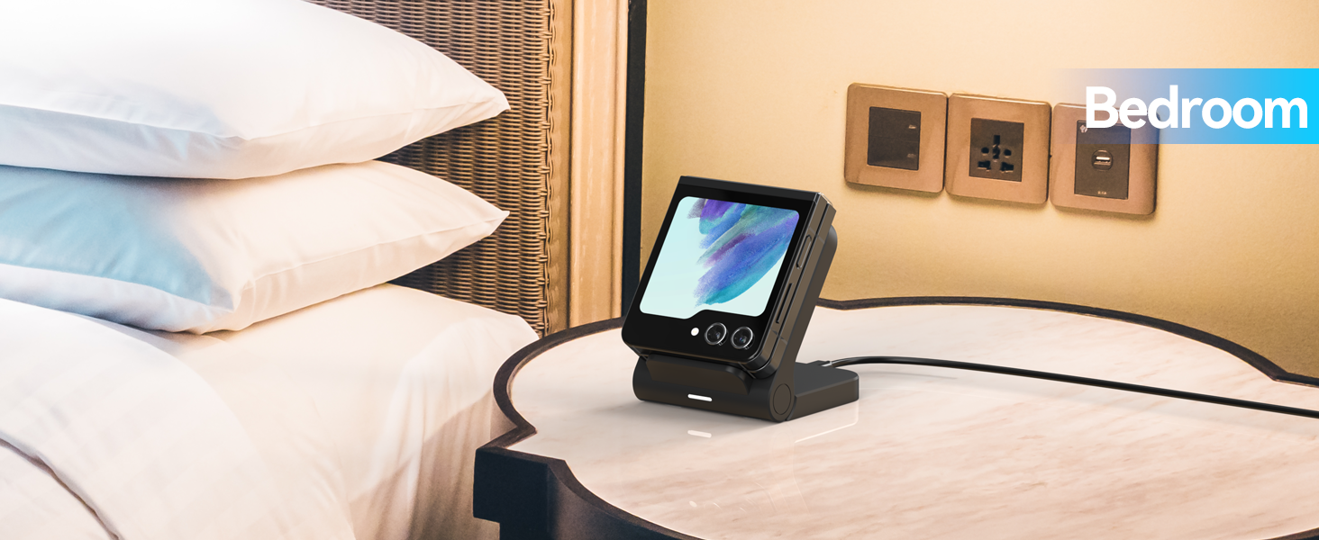 Black digital alarm clock on bedside table. Device has a large color display showing abstract blue and purple swirls. Two white pillows visible in background.