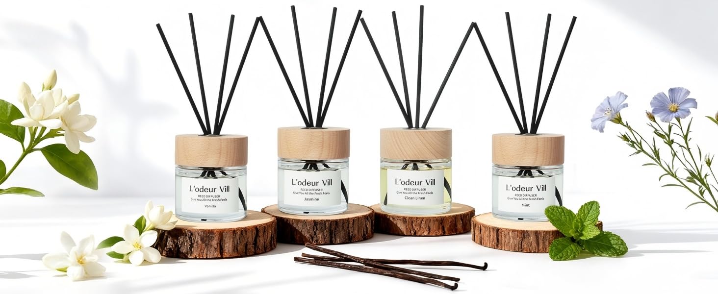 Set of four reed diffusers with black reeds displayed on wooden slice, surrounded by white flowers and greenery. Glass bottles contain fragrance oils.