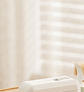 Partial view of a white table or shelf against a light-colored wall with vertical blinds visible in the background.