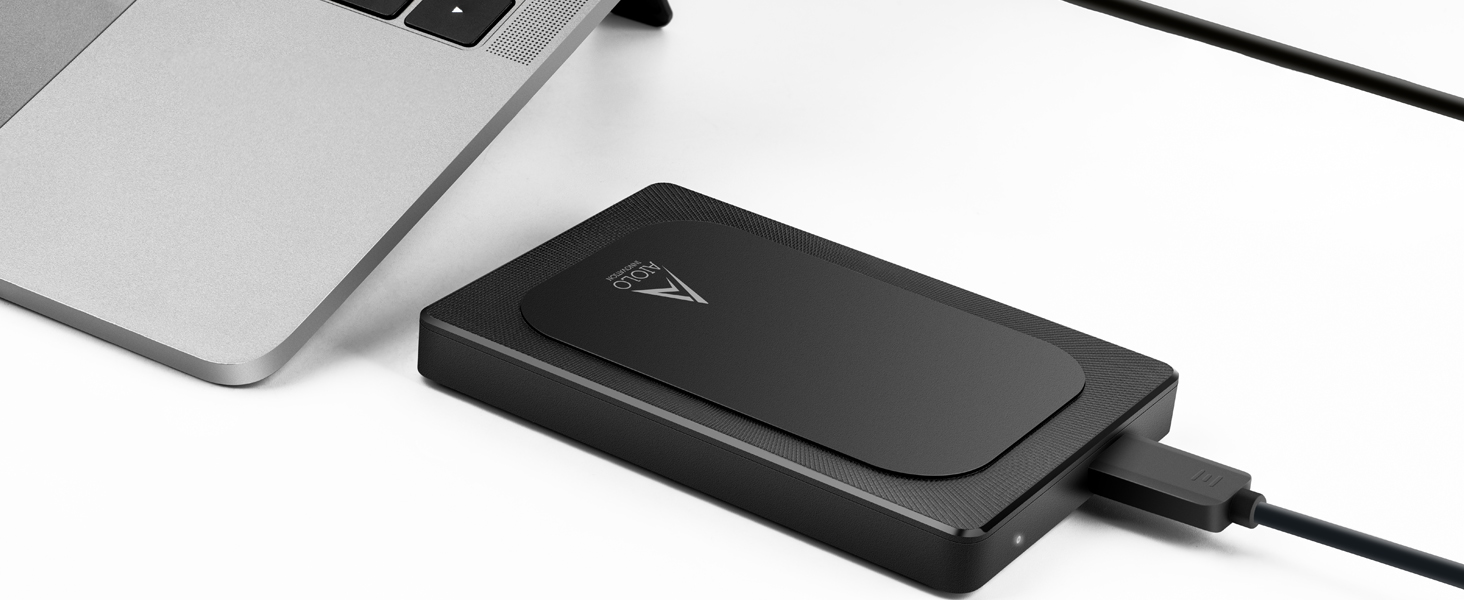 Black external hard drive connected to a laptop via USB cable. The device has a slim, rectangular design with rounded edges.