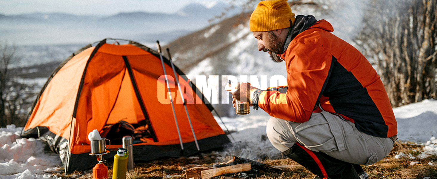 Person in orange jacket and yellow hat sitting by orange tent in snowy landscape, holding a camping mug.