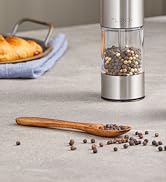 Stainless steel and glass pepper mill with whole peppercorns visible inside, shown on gray surface with wooden spoon and croissant in background.